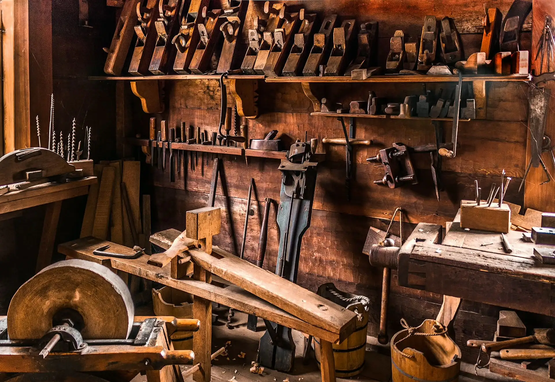 A traditional woodworking workshop — a wall of vintage hand planes, chisels, and tools above a workbench and grindstone
